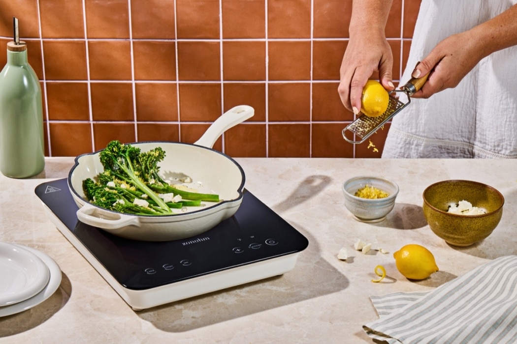 Person zesting a lemon over broccolini and feta cheese cooking in a cream enameled pan on a portable induction cooktop, styled with a terracotta tile backdrop