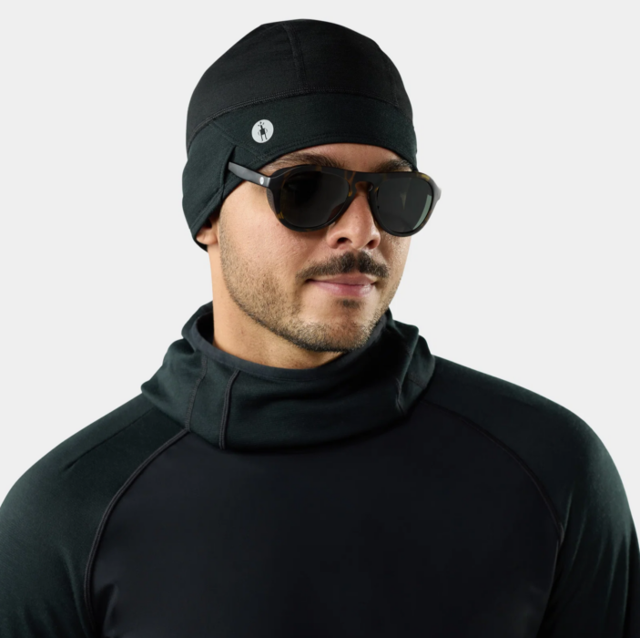 Male model wearing a black beanie, sunglasses and technical black outerwear against a white background.