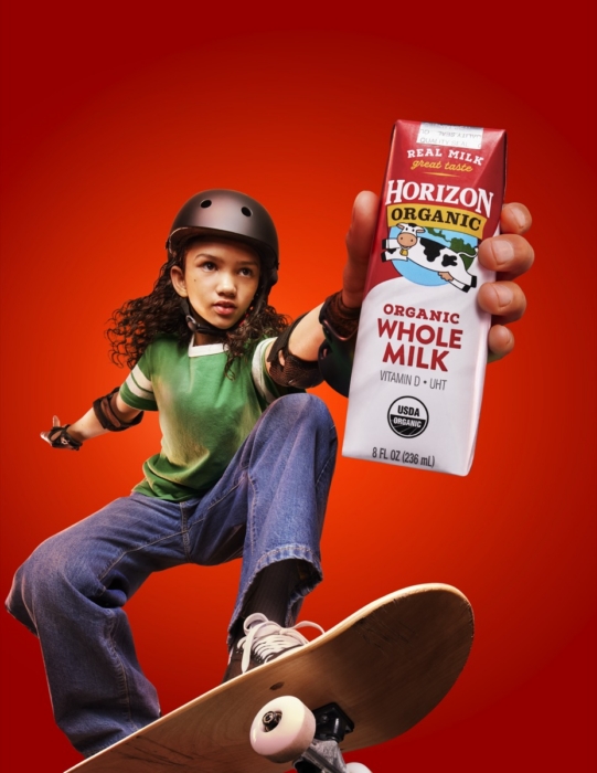 Young girl wearing a helmet and riding a skateboard while holding a Horizon Organic whole milk carton against a vibrant red studio background.