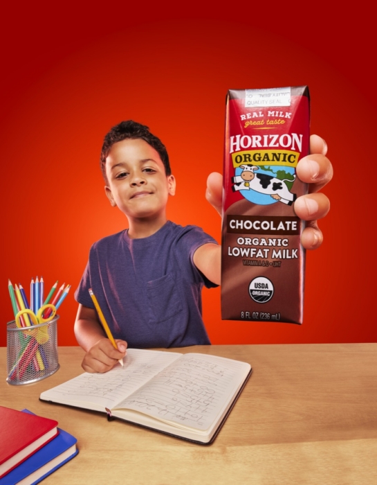 Smiling young student seated at a desk holding a Horizon Organic chocolate milk carton toward the camera against a bright red studio background.
